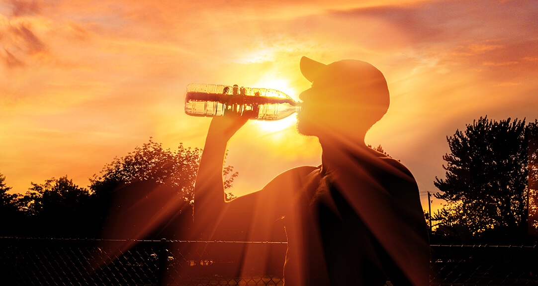 Silhouette of a person wearing a baseball cap drinking water from a plastic bottle, highlighting the importance of staying hydrated during heatwaves.