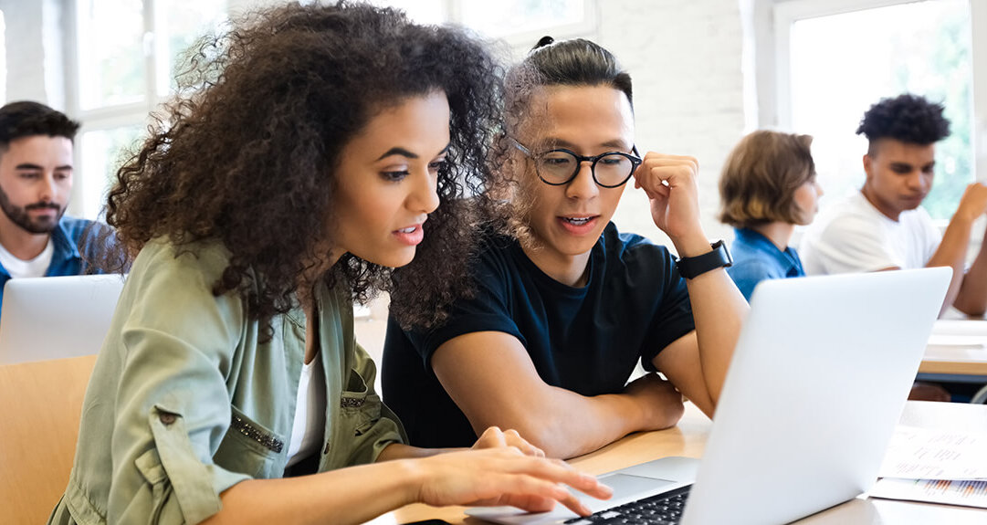 A young woman with curly hair and a young man with his hair pulled back are in a casual classroom setting both looking at a laptop.