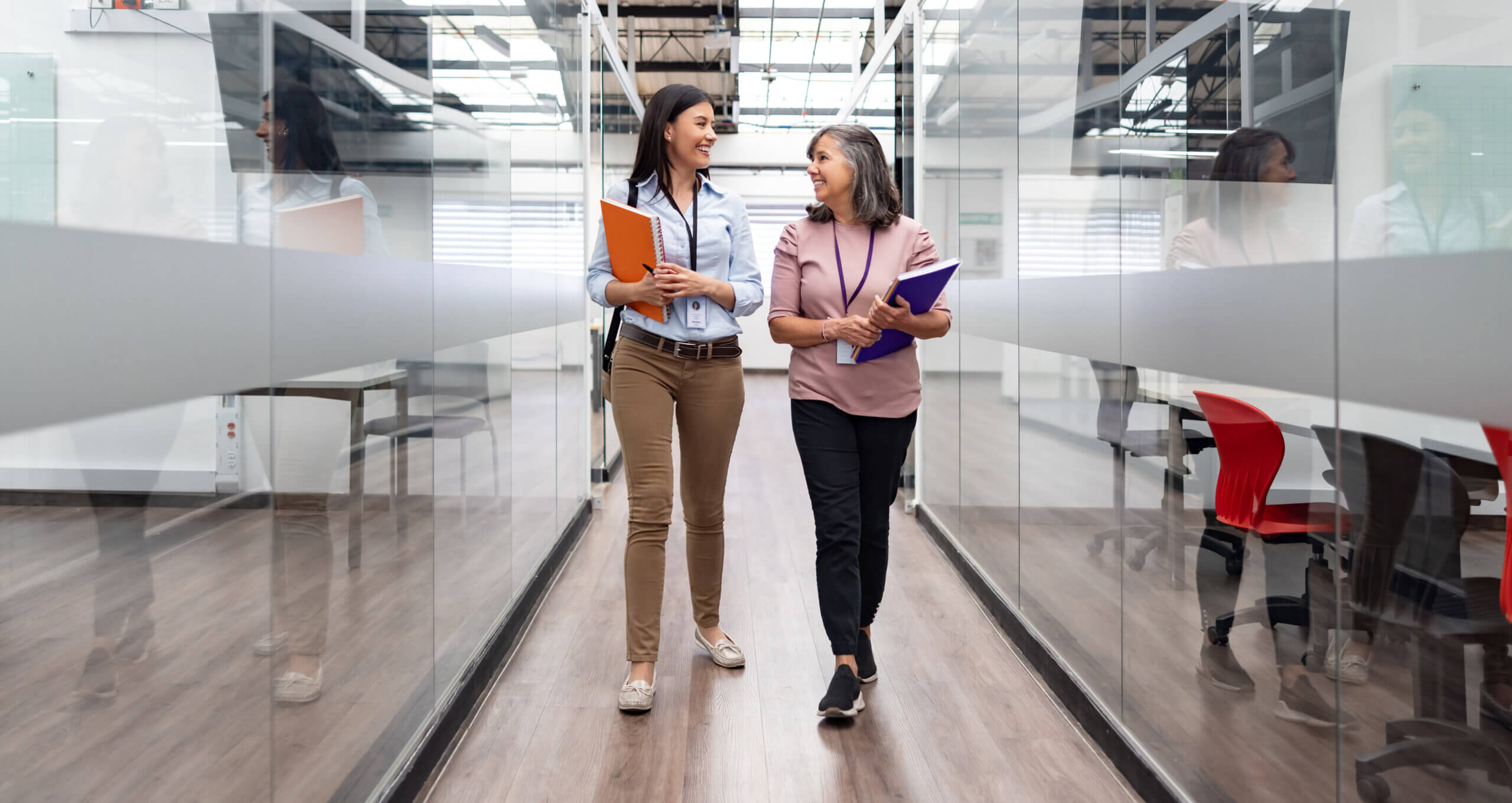 A pair of smiling female professors with books in their hands talk in the hallway of a university while walking toward a classroom.