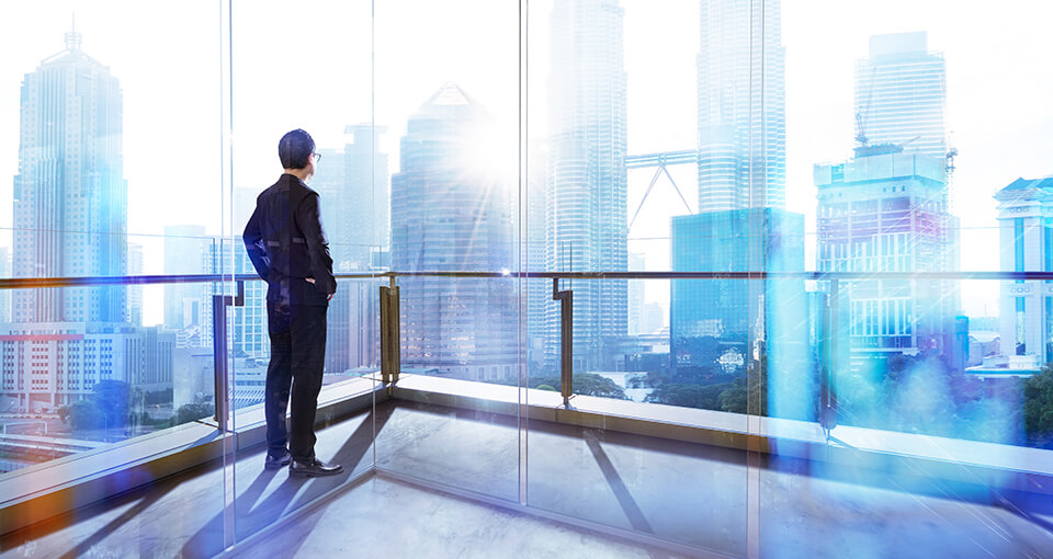 A business professional dressed in a suit stands on a balcony in a modern high-rise building. The person faces large glass windows that offer a sunlight view of a skyline filled with tall skyscrapers.