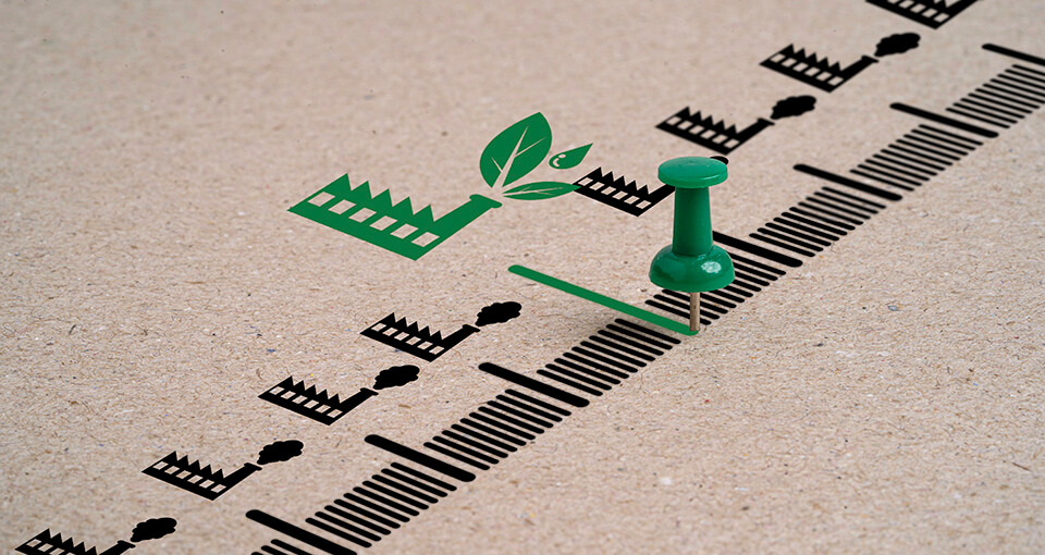 The image represents environmental sustainability, with a green pushpin placed along a timeline marked with black factory icons, each emitting pollution.