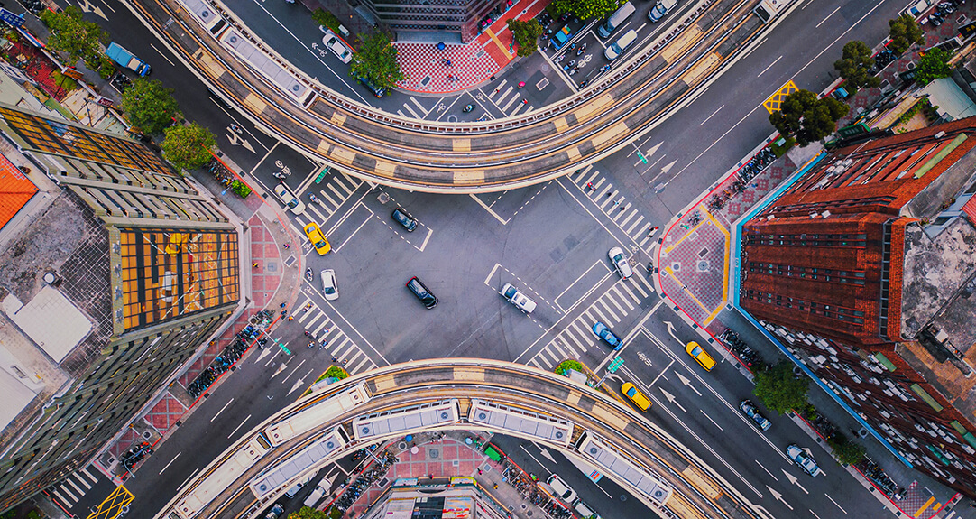 Aerial view of a vibrant urban scene at a large intersection. Several roads meet in the center, with cars moving in various directions, while elevated train tracks curve over the streets.