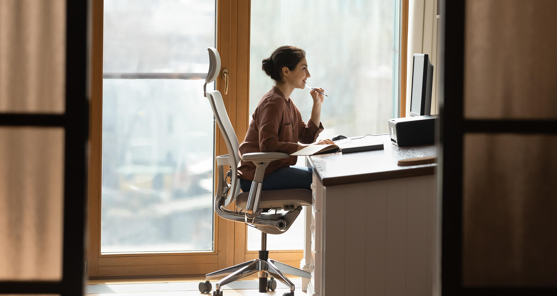 A ywoman is working in a modern home office, sitting at a desk with a computer and multiple notebooks. She’s focused, with her hand resting thoughtfully on her chin as she looks at the screen.