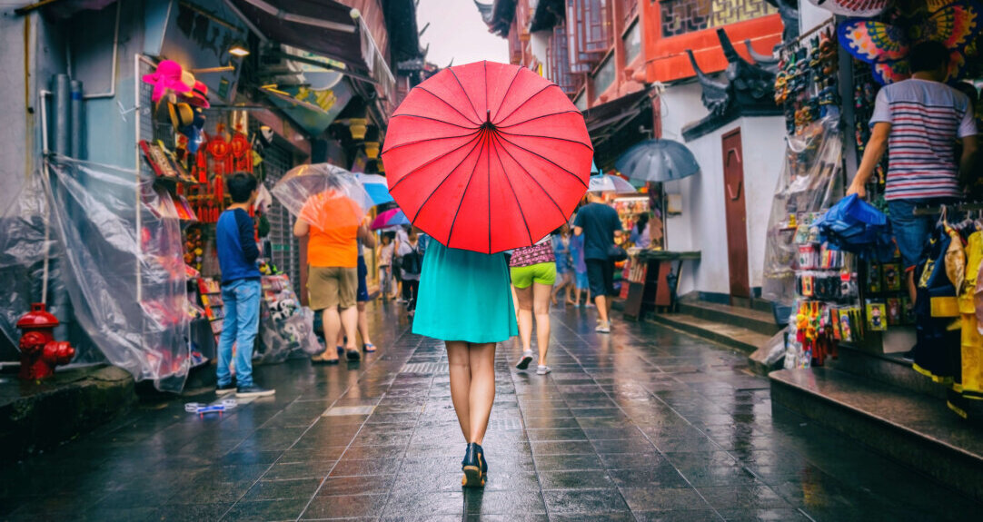 A woman holding a red umbrella stands in the center of the image with her back to the reader as she walks through a bustling Asian market street.