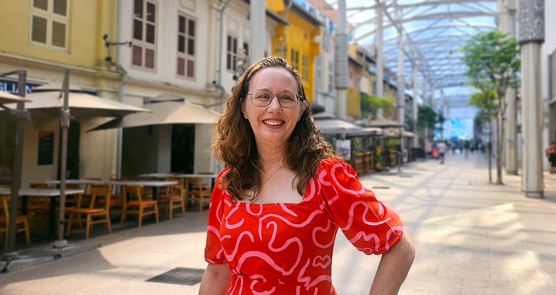 Jill Hoffman, regional head of internal audit for Munich Re Asia Pacific, Middle East and Africa, smiles while standing in a vibrant outdoor walkway with cafes and colorful buildings in the background.