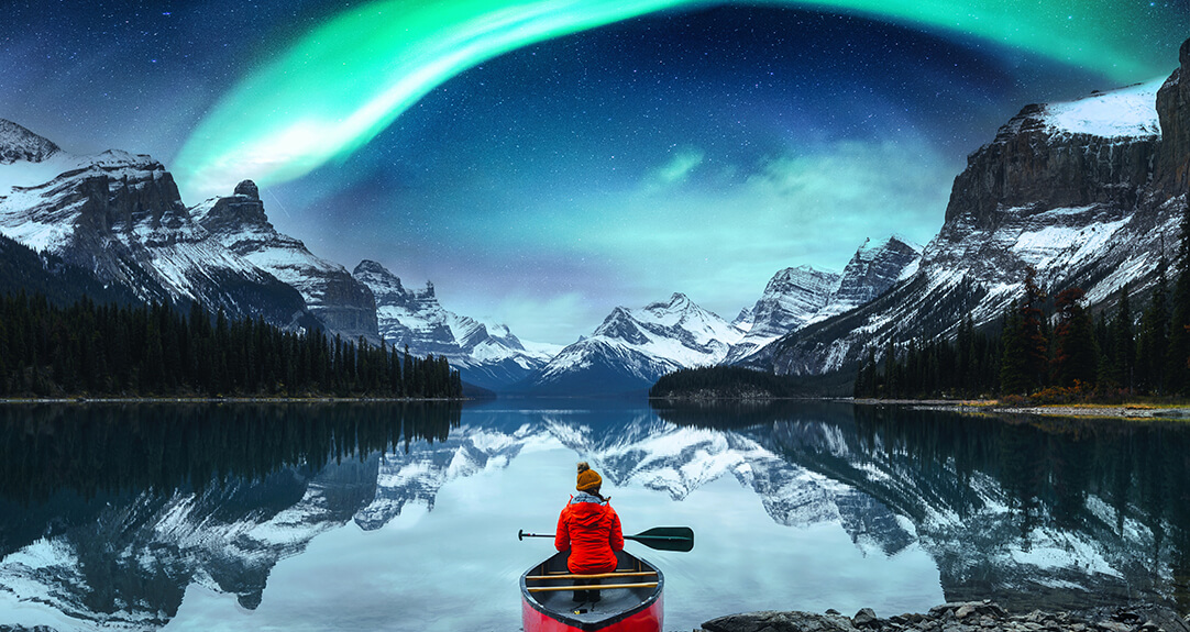 A person wearing a red jacket sits in a red canoe at the edge of a calm, mirror-like lake, surrounded by snow-capped mountains under a night sky featuring the aurora borealis.