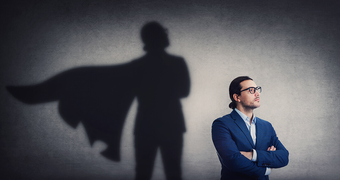 A confident man in a blue suit with arms crossed stands against a textured gray wall. Behind him, his shadow is cast as a superhero figure with a flowing cape, symbolizing inner strength and leadership potential.