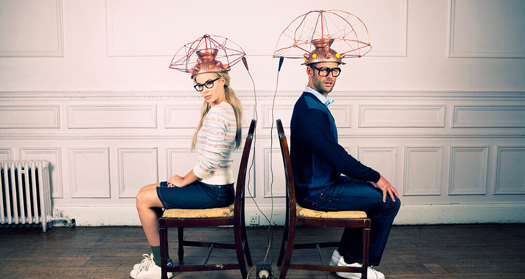 A man and woman sit back-to-back on wooden chairs in a vintage room, wearing quirky, retro-futuristic headgear with copper wires and a playful sci-fi vibe.